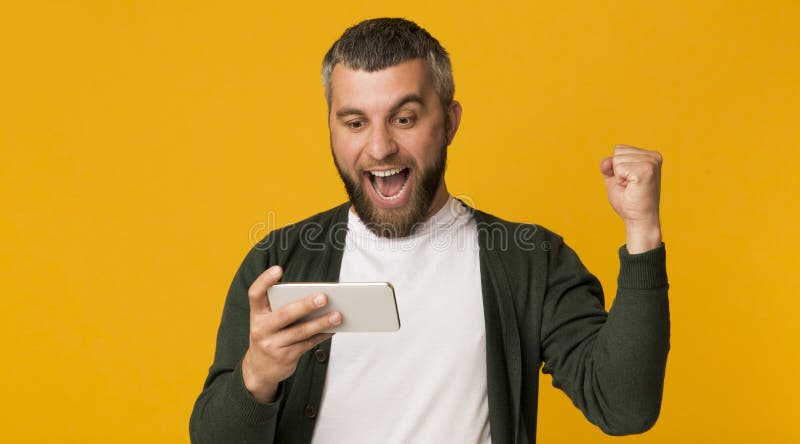 Эмоциональный бородатый мужчина, держащий смартфон и кулакивающий кулак стоковые фото