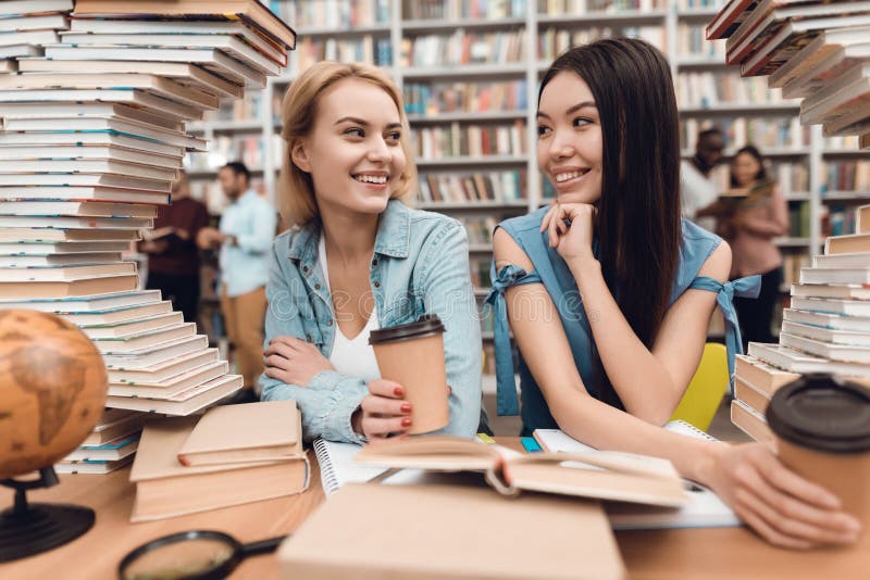 Этническая азиатская девушка и белая девушка окруженная книгами в библиотеке Студенты выпивают кофе стоковое изображение rf Этническая азиатская девушка и белая девушка окруженная книгами в библиотеке Студенты выпивают кофе стоковое изображение rf