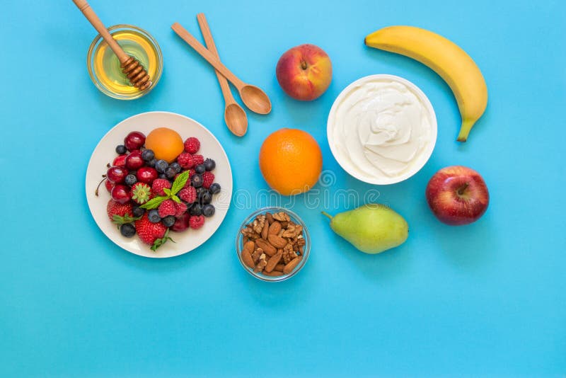 Greek yogurt around fruits orange, banana, pear, peach, apple, plate with strawberries, raspberries, blueberries and nuts, honey and 2 wooden spoons on light blue background. Horizontal. Top view. Greek yogurt around fruits orange, banana, pear, peach, apple, plate with strawberries, raspberries, blueberries and nuts, honey and 2 wooden spoons on light blue background. Horizontal. Top view.