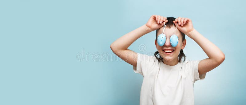 улыбающийся маленький мальчик держит пасхальные яйца в руке и закрывает ими глаза на синем фоне стоковые фотографии rf