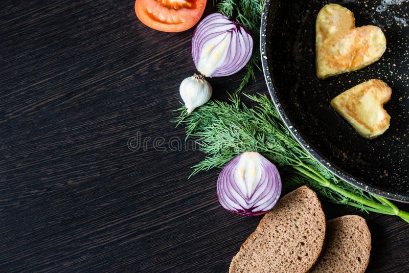 Яичницы в форме сердца в сковороде, около которой куски хлеба положения, красный лук, томат, чеснок и укроп стоковое изображение rf Яичницы в форме сердца в сковороде, около которой куски хлеба положения, красный лук, томат, чеснок и укроп стоковое изображение rf