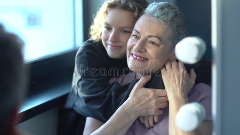 一个十几岁的女孩拥抱着她坐在镜子旁的灰发美丽的祖母. 两代一代 影视素材