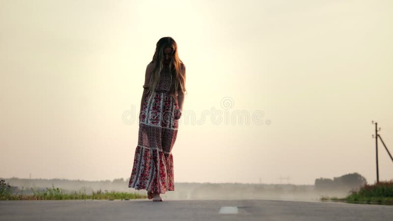 一个年轻漂亮的女孩在夏天温暖的夜晚赤脚穿着长裙在路上行走
