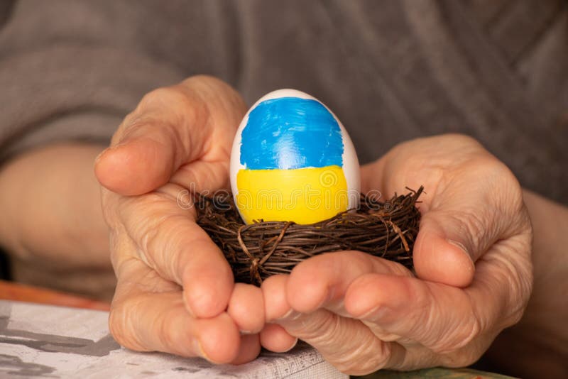 One white chicken egg with a painted flag of Ukraine in a straw basket in the hands of an old woman, culture and holidays 2022, stop the war. One white chicken egg with a painted flag of Ukraine in a straw basket in the hands of an old woman, culture and holidays 2022, stop the war