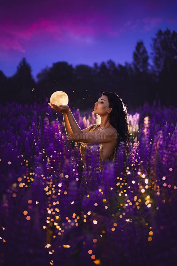 A brunette girl in a golden dress holding a moon in her hands and standing among a blooming purple lupine field with lights and fireflies. A magical night portrait. A brunette girl in a golden dress holding a moon in her hands and standing among a blooming purple lupine field with lights and fireflies. A magical night portrait.