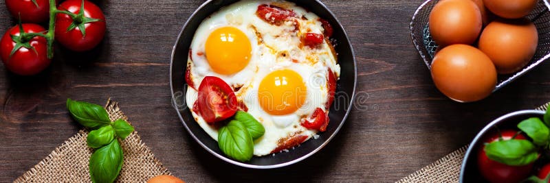 Delicious and healthy breakfast for one person: small pan with 2 fried eggs with fresh vegetables and cheese. Served on a dark wooden table with yellow napkin, fresh tomatoes and basil, brown bread. Banner. Delicious and healthy breakfast for one person: small pan with 2 fried eggs with fresh vegetables and cheese. Served on a dark wooden table with yellow napkin, fresh tomatoes and basil, brown bread. Banner