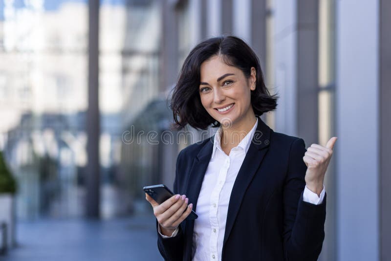 一位年轻成功女商人站在办公中心附近，手拿一部手机，手中竖着一根超级手指 免版税图库摄影