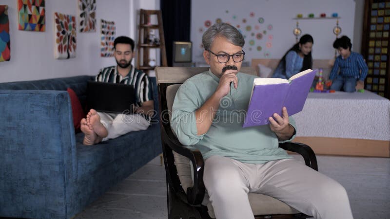一位年迈的祖父戴着眼镜，在家读一本印度三代家庭的书