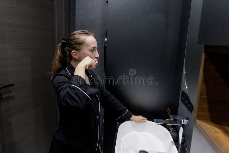 一位穿着舒适睡衣的女士在现代化的浴室里刷牙，享受着她每天的自我护理程序。 早晨例行活动 免版税库存图片