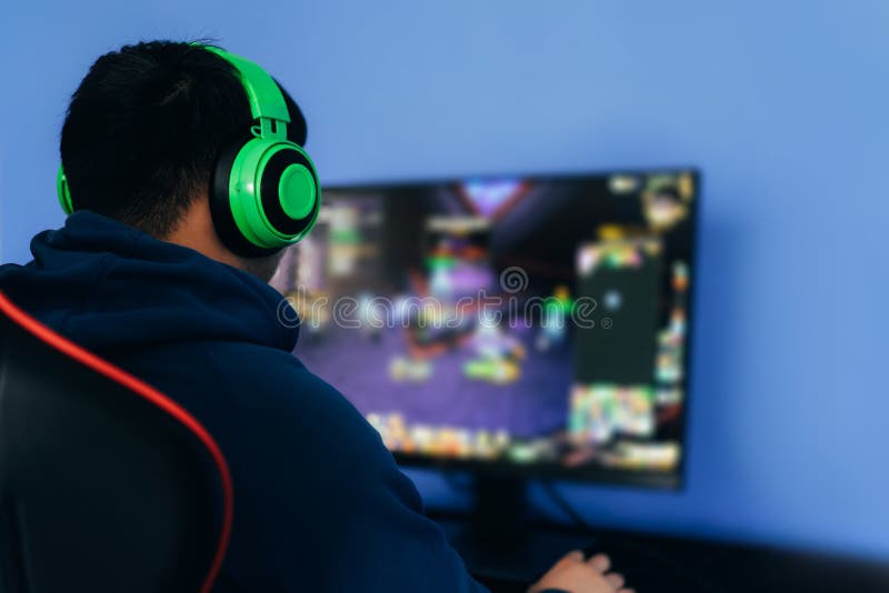 一名年轻男子在职业扶手椅上玩电脑游戏，戴着绿色耳机在显示器上玩直播流蓝色