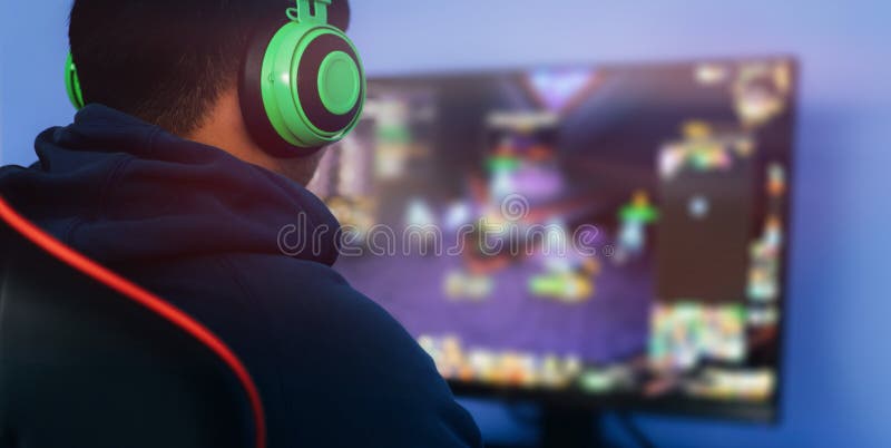一名年轻男子在职业扶手椅上玩电脑游戏，戴着绿色耳机在显示器上玩直播流蓝色