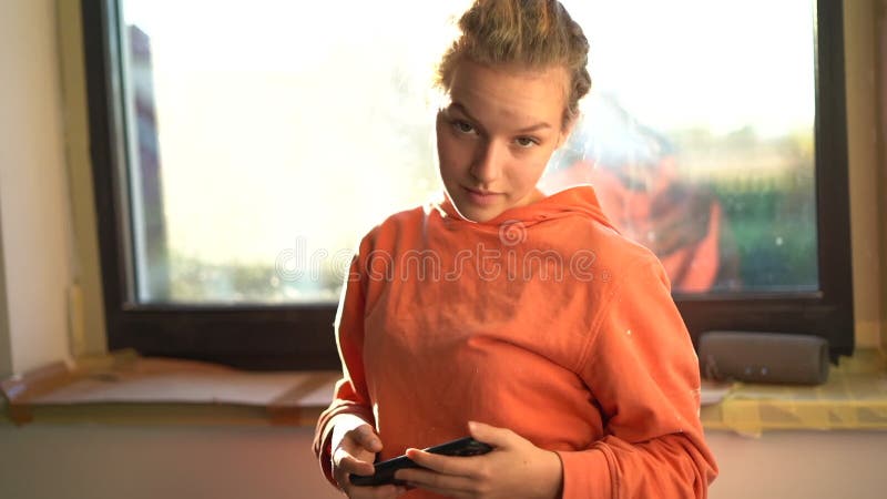 一头卷发的少年少女的欢快肖像,穿着一件橙色彩色连帽衫,在房子的窗前 股票录像