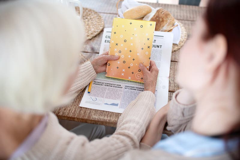 Reading news. Top view of grey-haired women reading news in the morning sitting near caregiver. Reading news. Top view of grey-haired women reading news in the morning sitting near caregiver