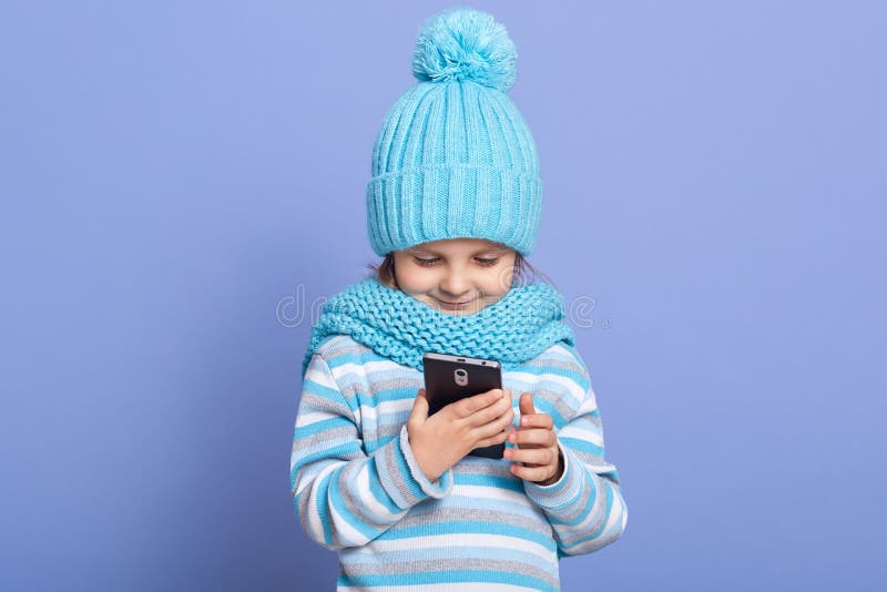 一幅细心可爱、头戴蓝帽子、围巾、条纹运动衫、站在淡紫色背景上的小女孩肖像 图库摄影 一幅细心可爱、头戴蓝帽子、围巾、条纹运动衫、站在淡紫色背景上的小女孩肖像 图库摄影