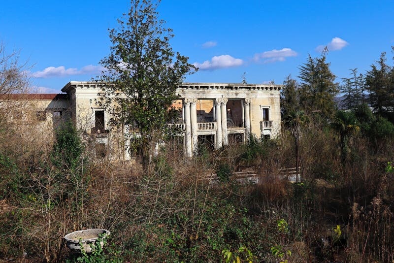 Abandoned, run-down and dilapidated large building, surrounded by lush vegetation of various trees and palms. The building no longer has a chance of a roof, only bare walls with remnants of ornaments remain, which tell about a once beautiful building full of life. The front entrance of the house has beautiful tall columns with decoration, small balconies with a nice balustrade. Windows and entrance doors are no longer in place. A decorative ledge can be seen at the top of the wall. Abandoned, run-down and dilapidated large building, surrounded by lush vegetation of various trees and palms. The building no longer has a chance of a roof, only bare walls with remnants of ornaments remain, which tell about a once beautiful building full of life. The front entrance of the house has beautiful tall columns with decoration, small balconies with a nice balustrade. Windows and entrance doors are no longer in place. A decorative ledge can be seen at the top of the wall