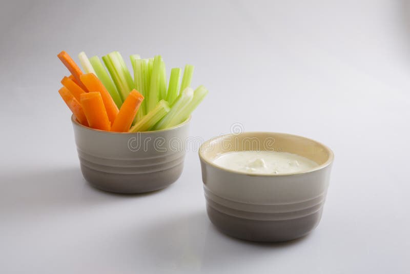 Close up isolated shot of a bowl of separated crunchy orange carrot slices and juicy green celery sticks with a white cup of blue cheese dipping sauce on a white background. Close up isolated shot of a bowl of separated crunchy orange carrot slices and juicy green celery sticks with a white cup of blue cheese dipping sauce on a white background