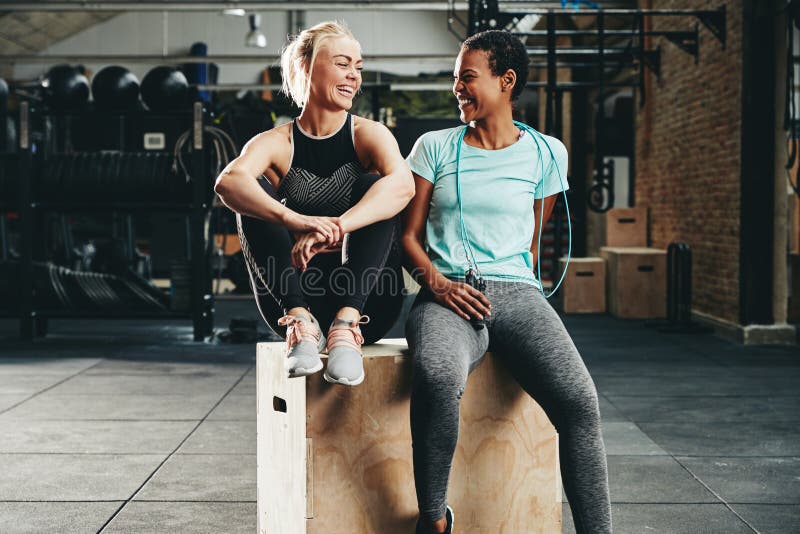 Two fit young female friends in sportswear laughing together while sitting on a box at the gym after a workout session. Two fit young female friends in sportswear laughing together while sitting on a box at the gym after a workout session