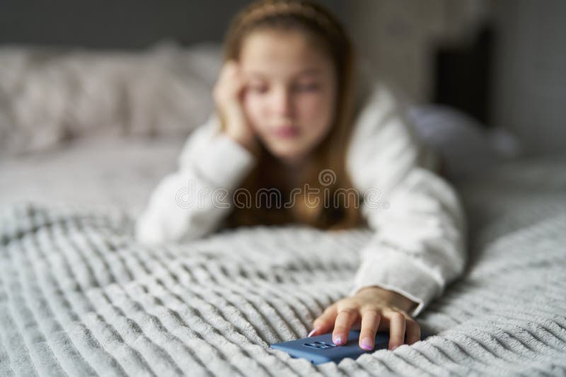 Unhappy Teenage Girl With Mobile Phone Lying On Bed At Home Anxious About Social Media Online Bullying And Using Phone Too Much With Focus On Foreground. Unhappy Teenage Girl With Mobile Phone Lying On Bed At Home Anxious About Social Media Online Bullying And Using Phone Too Much With Focus On Foreground