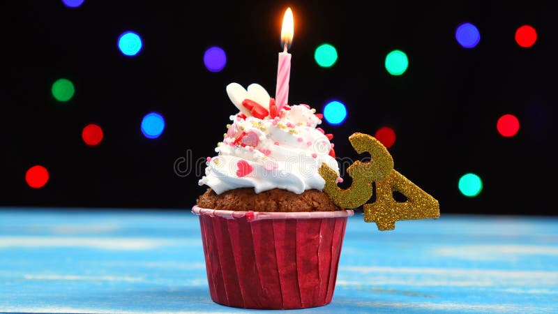 与燃烧的蜡烛和第34的可口生日杯形蛋糕在多彩多姿的被弄脏的光背景