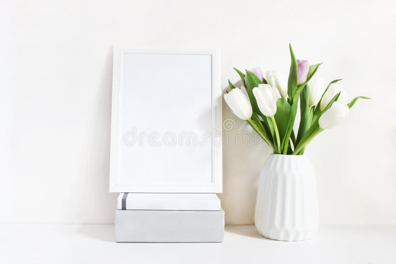 White blank wooden frame mockup with spring bouquet of tulips in porcelain vase and pile of books lying on the table. Poster product design, tyled stock feminine photography. Home decor. White blank wooden frame mockup with spring bouquet of tulips in porcelain vase and pile of books lying on the table. Poster product design, tyled stock feminine photography. Home decor.