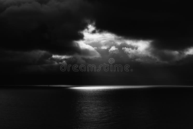 Abstract black and white image of dark storm clouds over dark ocean with one spot of light beaming through. Abstract black and white image of dark storm clouds over dark ocean with one spot of light beaming through