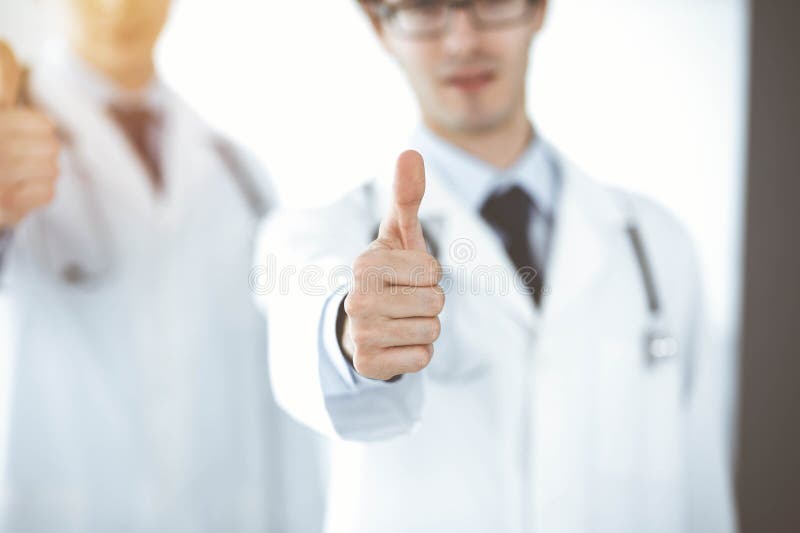 两位男医生站在一起，竖起大拇指，象征着在阳光明媚的诊所为病人提供最佳服务 图库摄影