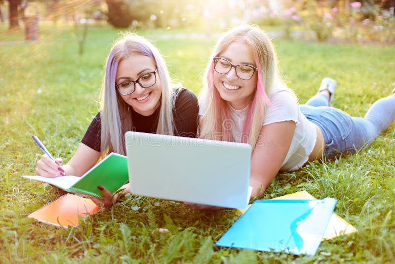两名千禧一代的大学生女孩躺在校园里的草地上，年轻的微笑朋友们用笔记本电脑，用小册子写作 图库摄影