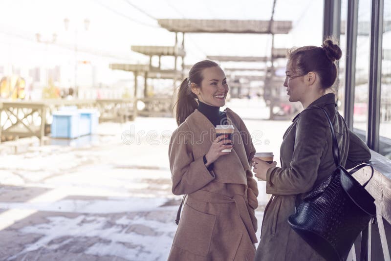 两名年轻时髦的妇女外套的和用咖啡手谈话在城市街道上，用餐步行，无忧无虑