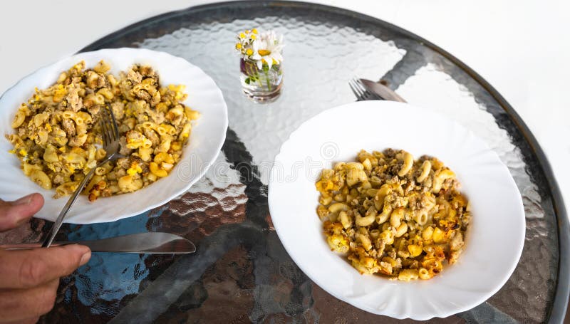 Baked pasta with minced meat and beaten egg in two plate on circle glass table outdoor. Part of hand holding cutlery. Baked pasta with minced meat and beaten egg in two plate on circle glass table outdoor. Part of hand holding cutlery