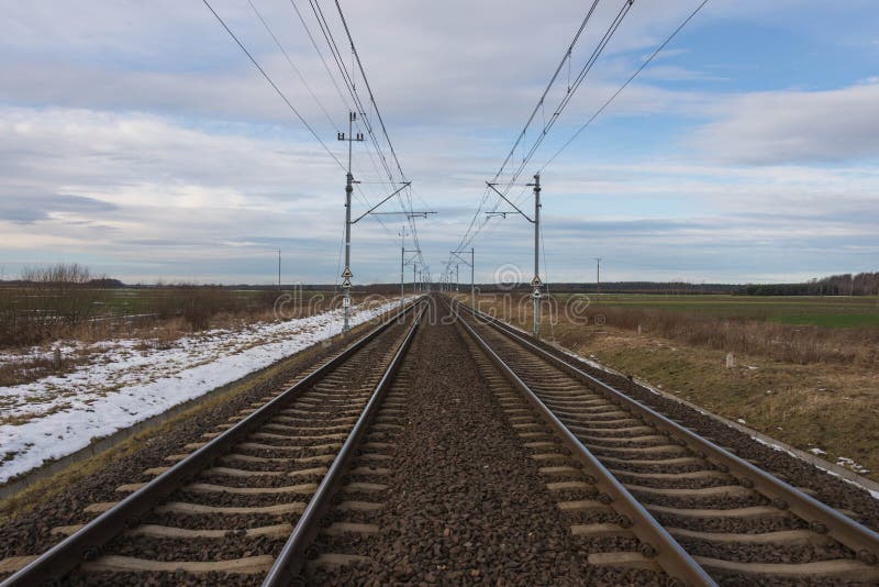 A view of the two-track, electrified railway track.
Winter, cloudy day. The ground is covered with sheets of snow. A two-track, electrified railway track. You can see the elements of electric traction in the form of metal poles, electric cables. Straight rails leading to the horizon. On the embankment you can see patches of a thin layer of snow. There are fields and meadows on both sides of the tracks. A view of the two-track, electrified railway track.
Winter, cloudy day. The ground is covered with sheets of snow. A two-track, electrified railway track. You can see the elements of electric traction in the form of metal poles, electric cables. Straight rails leading to the horizon. On the embankment you can see patches of a thin layer of snow. There are fields and meadows on both sides of the tracks.