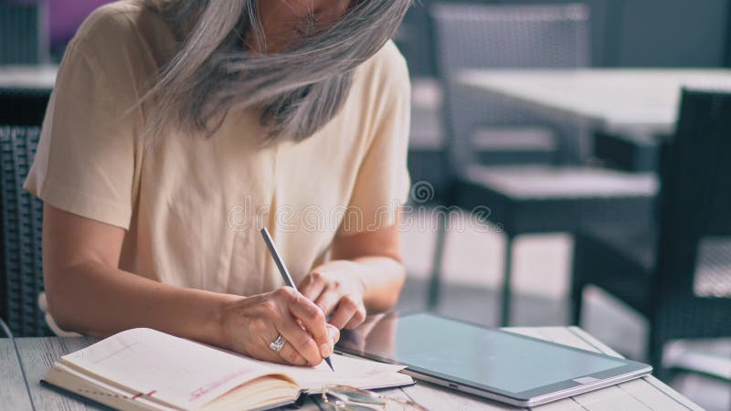 中年悦目亚裔妇女在咖啡馆坐并且做在她的笔记本的笔记