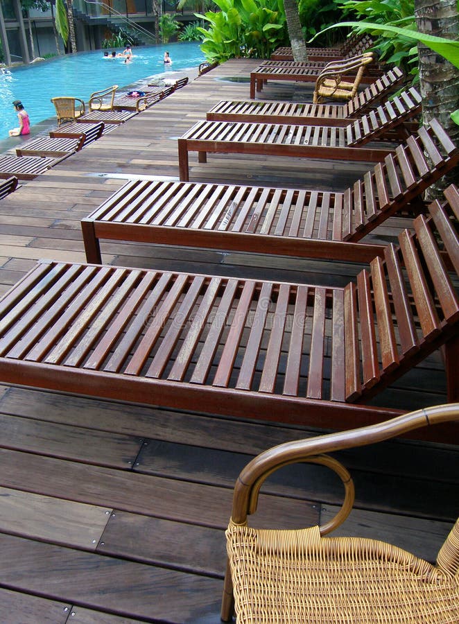 A row of deckchairs or lounge chairs by a pool side of a tropical island seaside resort. Samples of comfortable outdoor furniture placed on wooden board deck and green fresh landscaping around the swimming pool. Relaxing vacation mood. A row of deckchairs or lounge chairs by a pool side of a tropical island seaside resort. Samples of comfortable outdoor furniture placed on wooden board deck and green fresh landscaping around the swimming pool. Relaxing vacation mood.