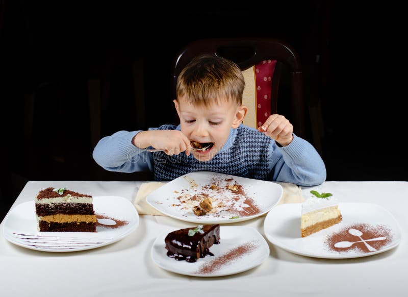 Cute little boy enjoying treat party cakes hungrily gulping down a big mouthful with slices variety different cakes displayed in front of him. Cute little boy enjoying treat party cakes hungrily gulping down a big mouthful with slices variety different cakes displayed in front of him