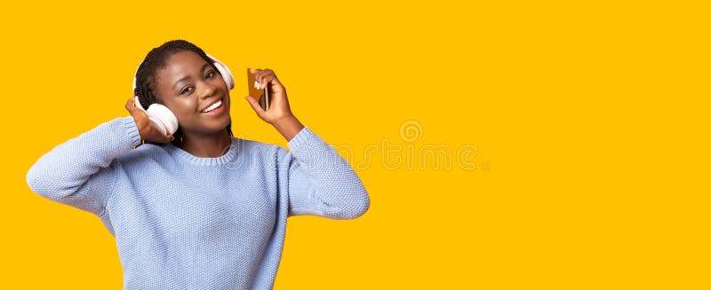 Happy african girl enjoying new music application for smartphone over yellow background, panorama with free space. Happy african girl enjoying new music application for smartphone over yellow background, panorama with free space