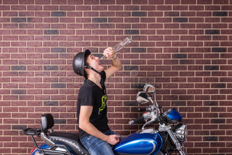 Young man sitting on a motorbike in his helmet drinking alcohol directly from the bottle as he satisfies hid addiction, copyspace on a brick wall. Young man sitting on a motorbike in his helmet drinking alcohol directly from the bottle as he satisfies hid addiction, copyspace on a brick wall