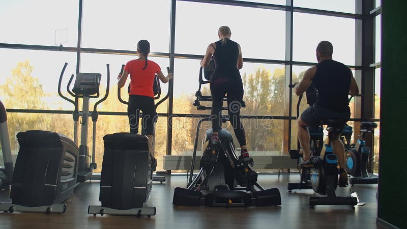 从后面看：在健身房的有氧区，女性使用椭圆机，男性使用健身车