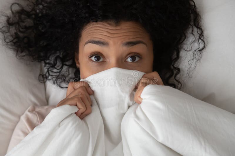 Top view close up of millennial african American girl lying in bed covered with white sheets look at camera, black young woman peer out of warm fluffy duvet, relaxing waking up in bedroom. Top view close up of millennial african American girl lying in bed covered with white sheets look at camera, black young woman peer out of warm fluffy duvet, relaxing waking up in bedroom