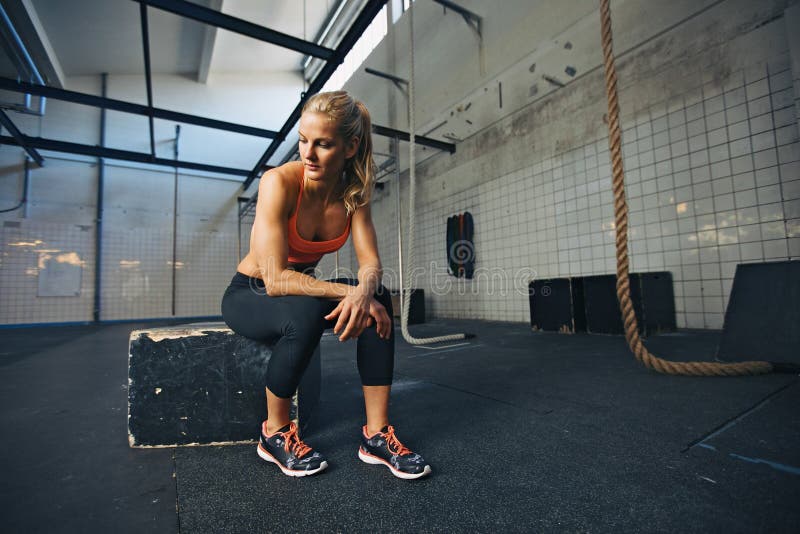 Caucasian female athlete sitting on a box at gym thinking. Fit young woman taking break after her workout at gym. Caucasian female athlete sitting on a box at gym thinking. Fit young woman taking break after her workout at gym.