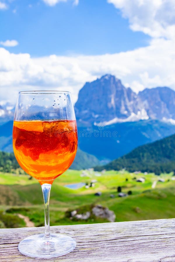 View of the traditional Italian alcoholic drink Aperol Spritz on the background of colorful Italian meadows and the Dolomites Alps mountains. village St. Cristina di Val Gardena Bolzano Seceda, Italy. View of the traditional Italian alcoholic drink Aperol Spritz on the background of colorful Italian meadows and the Dolomites Alps mountains. village St. Cristina di Val Gardena Bolzano Seceda, Italy