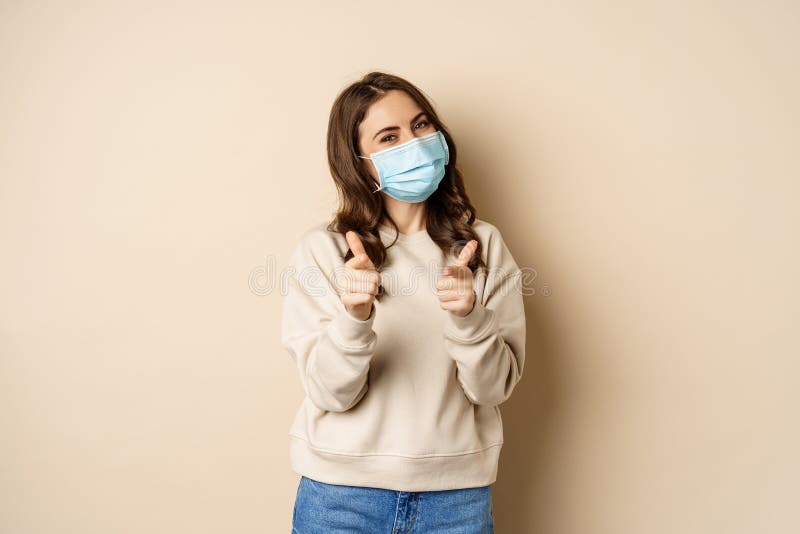 Hey you congrats. Smiling beautiful woman in medical face mask, pointing fingers at camera, standing over beige studio background. Hey you congrats. Smiling beautiful woman in medical face mask, pointing fingers at camera, standing over beige studio background.