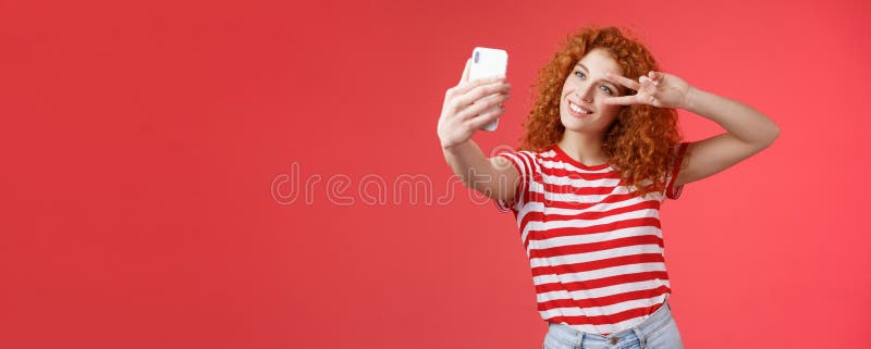 Hey followers how your summer holidays. Cheerful self-assured stylish fashionable redhead curly girl strike pose show peace victory gesture look smartphone screen taking selfie red background. Hey followers how your summer holidays. Cheerful self-assured stylish fashionable redhead curly girl strike pose show peace victory gesture look smartphone screen taking selfie red background.