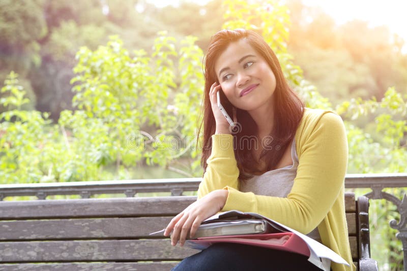 Beautiful young female university student with books and files on lap, using her phone, sitting on wooden bench in a park. Beautiful young female university student with books and files on lap, using her phone, sitting on wooden bench in a park.