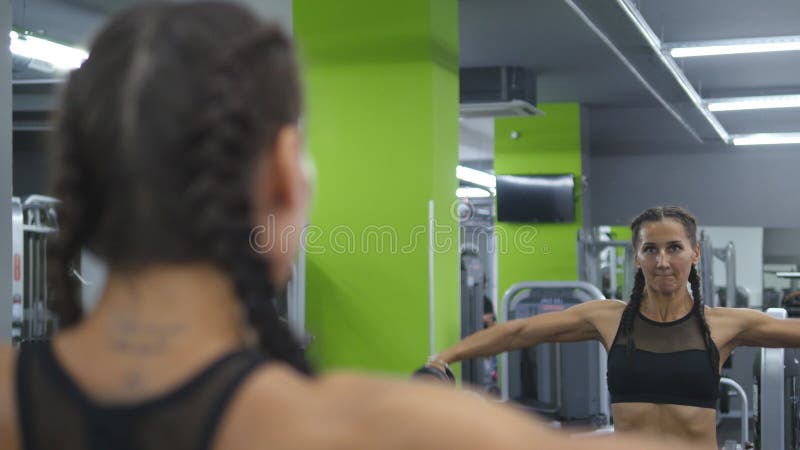 年轻俏丽的妇女在举和降低在镜子前面的健身房训练哑铃 体育健身女孩