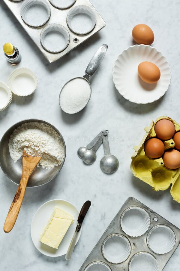 The ingredients for baking muffins or cupcakes are laid out on a marble countertop. This top down shot shows cupcake or muffin tins with papers, flour in a metal bowl with a wooden spoon, sugar in a measuring cup, vanilla, measuring spoons, a dish of butter with a knife, and a half dozen eggs. The ingredients for baking muffins or cupcakes are laid out on a marble countertop. This top down shot shows cupcake or muffin tins with papers, flour in a metal bowl with a wooden spoon, sugar in a measuring cup, vanilla, measuring spoons, a dish of butter with a knife, and a half dozen eggs.