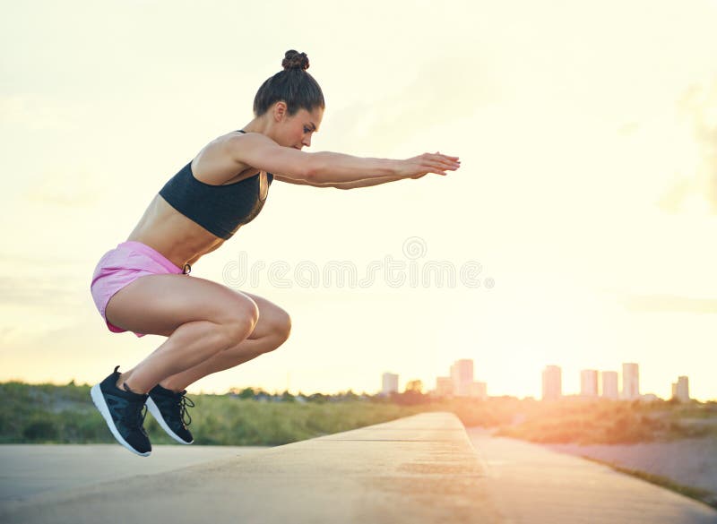 Healthy fit young woman doing crossfit exercises leaping on and off a low stone wall at the side of a promenade with the rising sun and cityscape in the distance. Healthy fit young woman doing crossfit exercises leaping on and off a low stone wall at the side of a promenade with the rising sun and cityscape in the distance