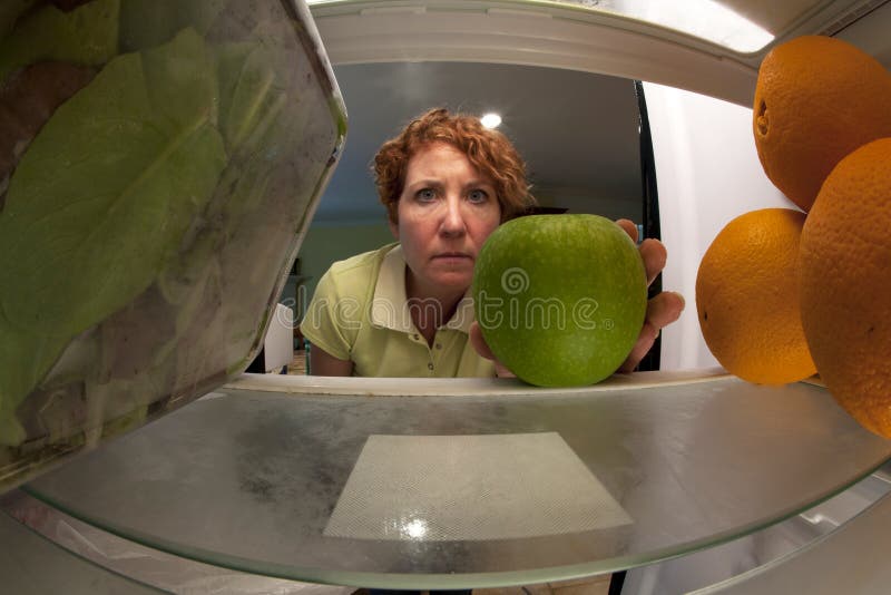 View from inside refrigerator of a woman selecting an apple surrounded by salad and oranges. View from inside refrigerator of a woman selecting an apple surrounded by salad and oranges