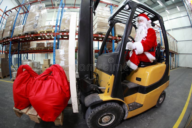 Santa Claus as a forklift operator at work in warehouse. 2 large red sack at the front of forklift.fish-eye lens.profile view. Santa Claus as a forklift operator at work in warehouse. 2 large red sack at the front of forklift.fish-eye lens.profile view.