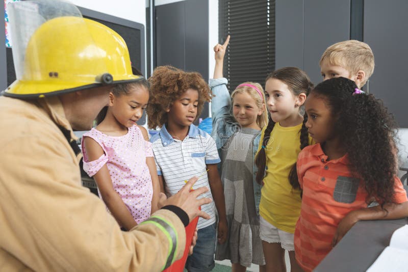 Rear view of a Caucasian firefighter wearing a fire helmet over his head and teaching about fire extinguisher to school kids in classroom at school. Rear view of a Caucasian firefighter wearing a fire helmet over his head and teaching about fire extinguisher to school kids in classroom at school