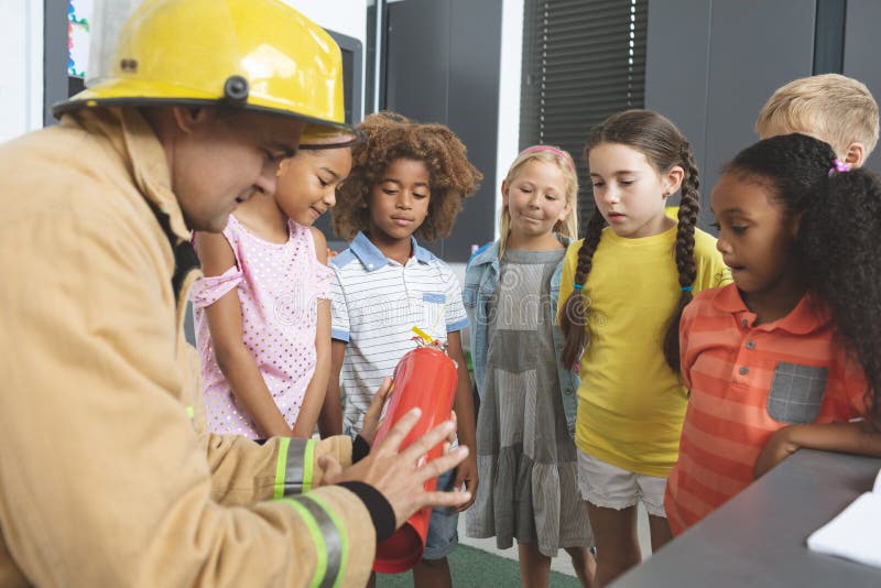 Rear view of a Caucasian firefighter wearing a fire helmet over his head and teaching about fire extinguisher to school kids in classroom at school. Rear view of a Caucasian firefighter wearing a fire helmet over his head and teaching about fire extinguisher to school kids in classroom at school