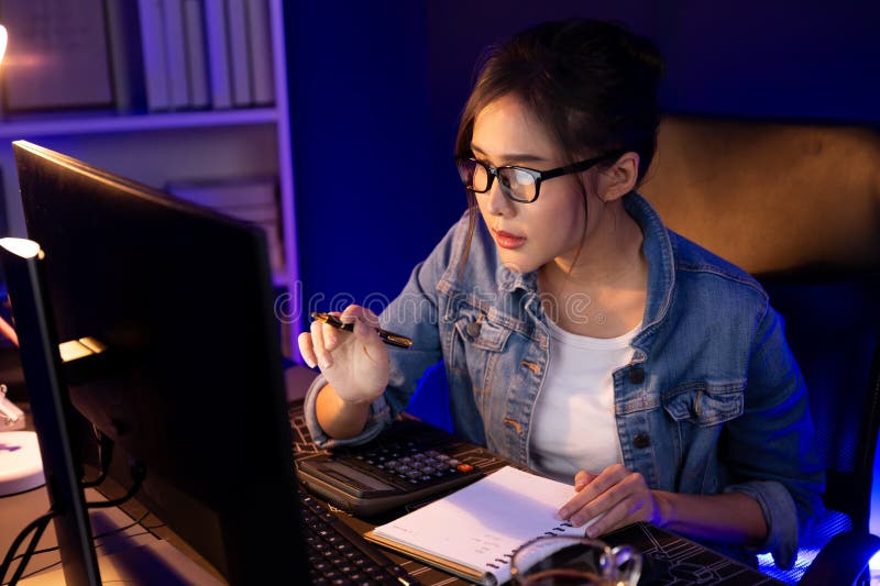 Thinking young beautiful Asian creative woman looking on pc with taking note on paper for memo in sales target report on business market graph report at neon light modern office at night. Stratagem. Thinking young beautiful Asian creative woman looking on pc with taking note on paper for memo in sales target report on business market graph report at neon light modern office at night. Stratagem.